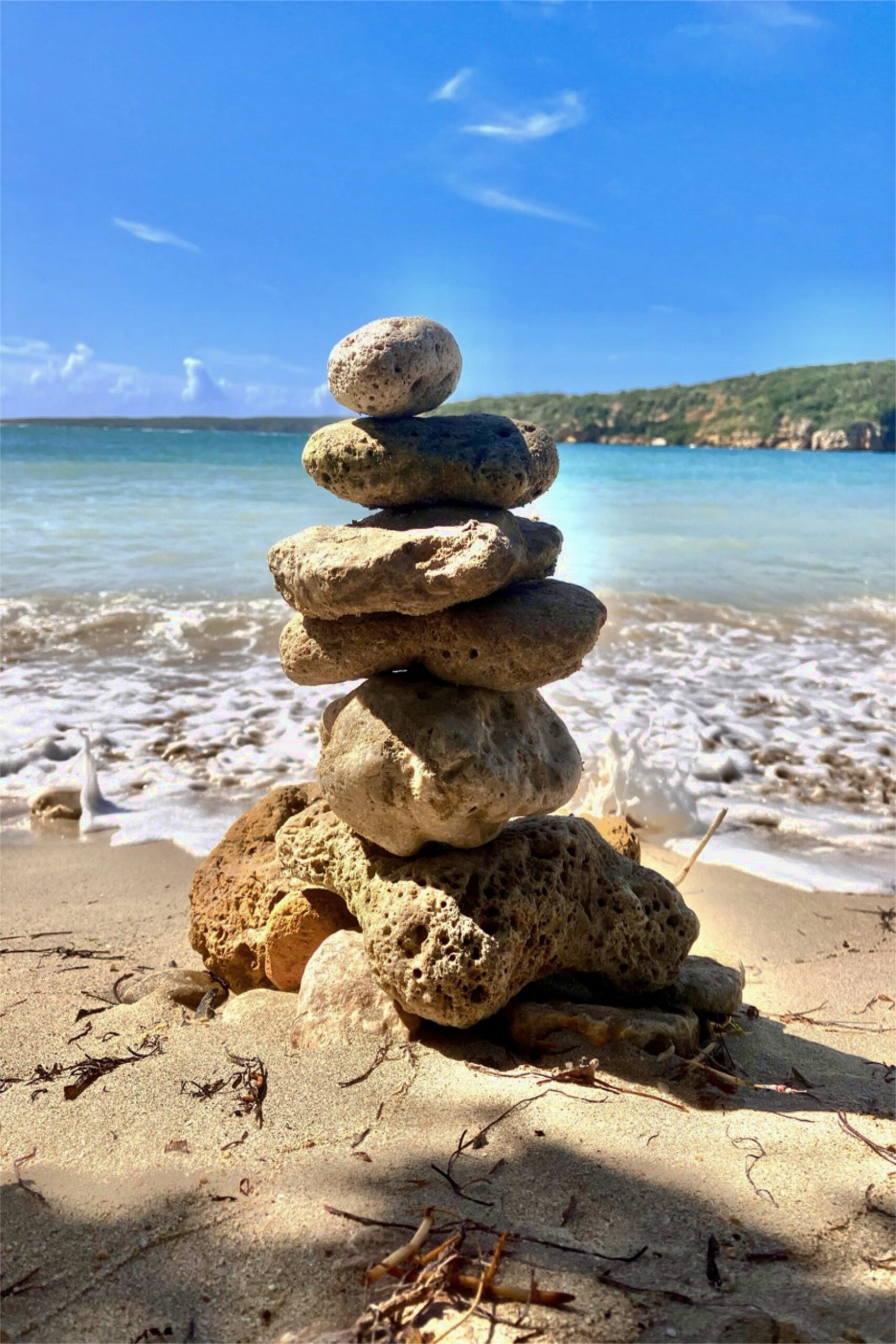 Piedras apiladas en equilibrio sobre la arena, frente al mar y bajo un cielo azul despejado.
