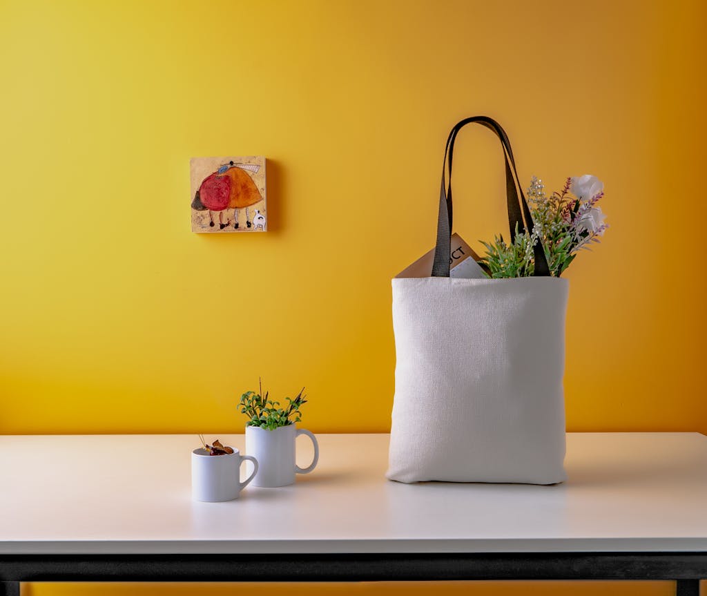 Bolsa de tela blanca sobre una mesa frente a una pared amarilla, con plantas pequeñas y algunos objetos cotidianos en un espacio doméstico sencillo.