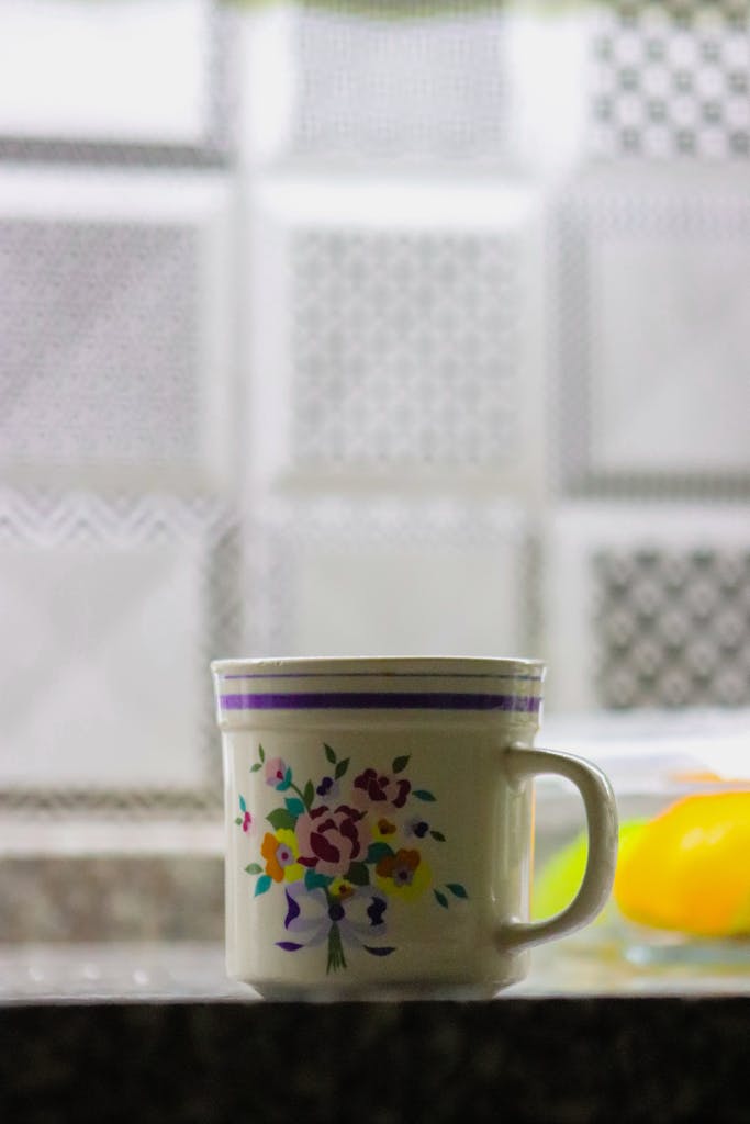 Taza con flores sobre el borde de una mesa, frente a una ventana con luz suave, en un rincón doméstico tranquilo y sencillo.