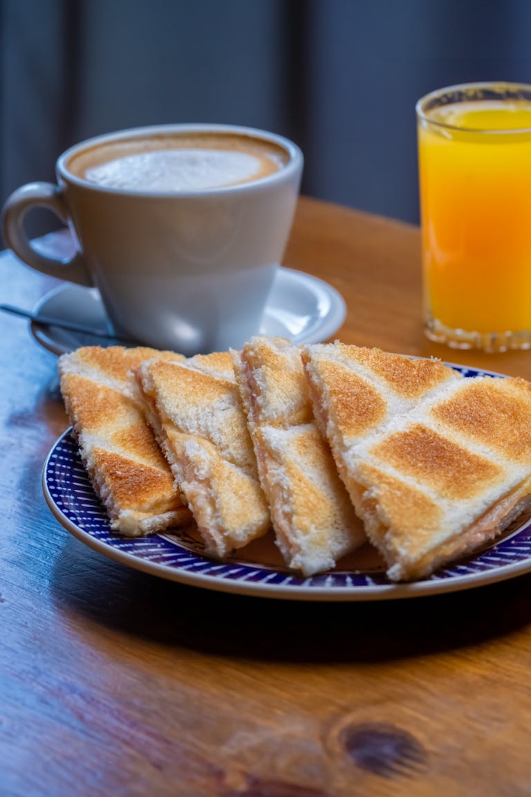 Desayuno delicioso con sándwiches tostados, cappuccino y jugo de naranja sobre una mesa de madera.