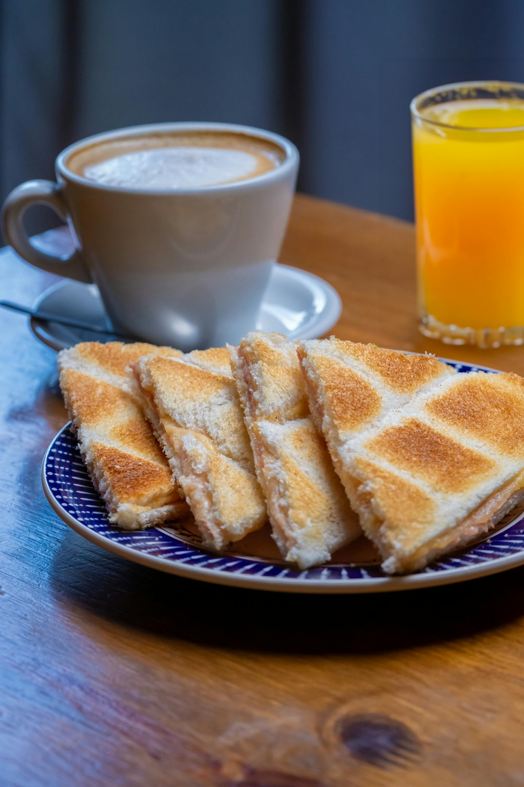 Desayuno delicioso con sándwiches tostados, cappuccino y jugo de naranja sobre una mesa de madera.
