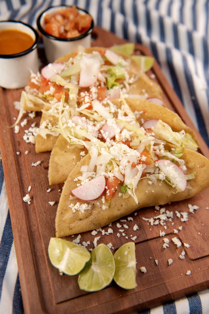 Tacos dorados de papa servidos sobre una tabla de madera, con lechuga, queso fresco, salsa, crema, rábanos y limones.