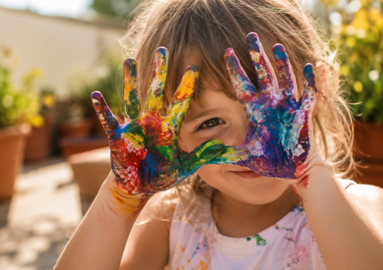 Niña con las manos cubiertas de pintura de colores durante actividades creativas de verano al aire libre.