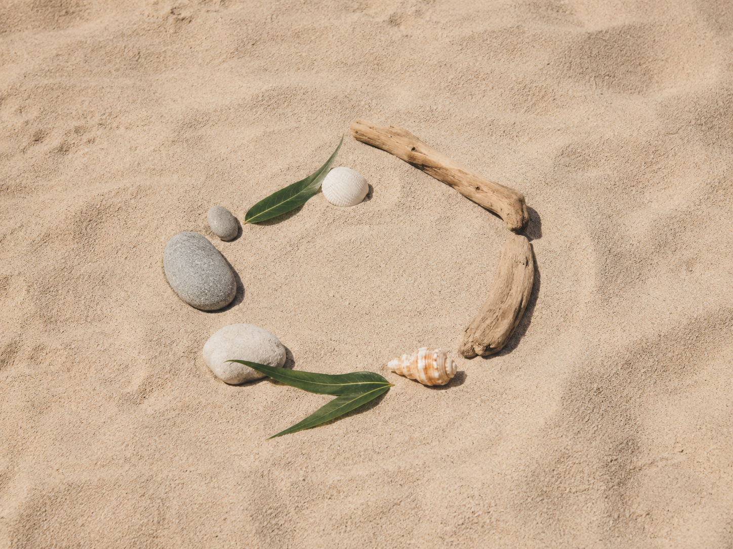 Composición sencilla de piedras, hojas, conchas y ramas sobre la arena para una actividad creativa de verano.