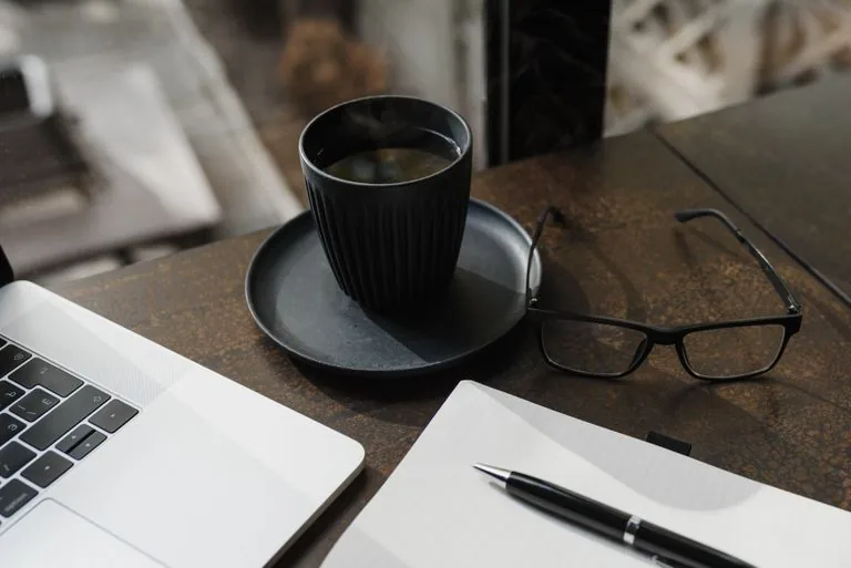 **Texto alternativo:** Mesa con una laptop, una taza de café, lentes y una libreta abierta junto a un bolígrafo.